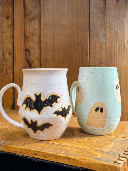Two handmade ceramic mugs, one with a bat design and the other with a ghost design, both with speckled backgrounds.