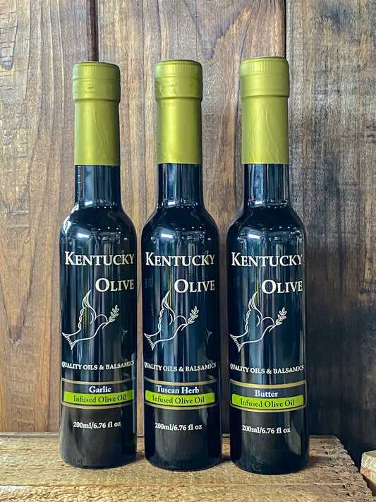 Three bottles of Kentucky Olive garlic infused olive oil, placed upright on a surface with a wooden background.