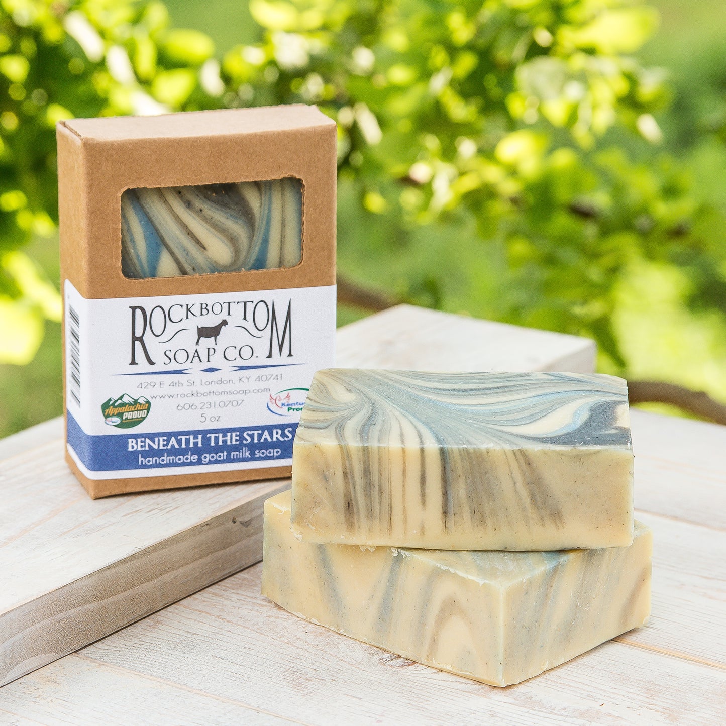 Swirled blue and black goat milk soap bar on a handmade pottery soap dish with green foliage background – fruity and vibrant artisan soap