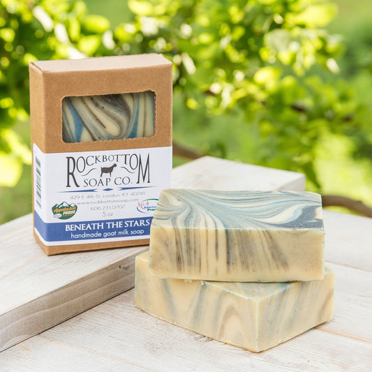 Swirled blue and black goat milk soap bar on a handmade pottery soap dish with green foliage background – fruity and vibrant artisan soap