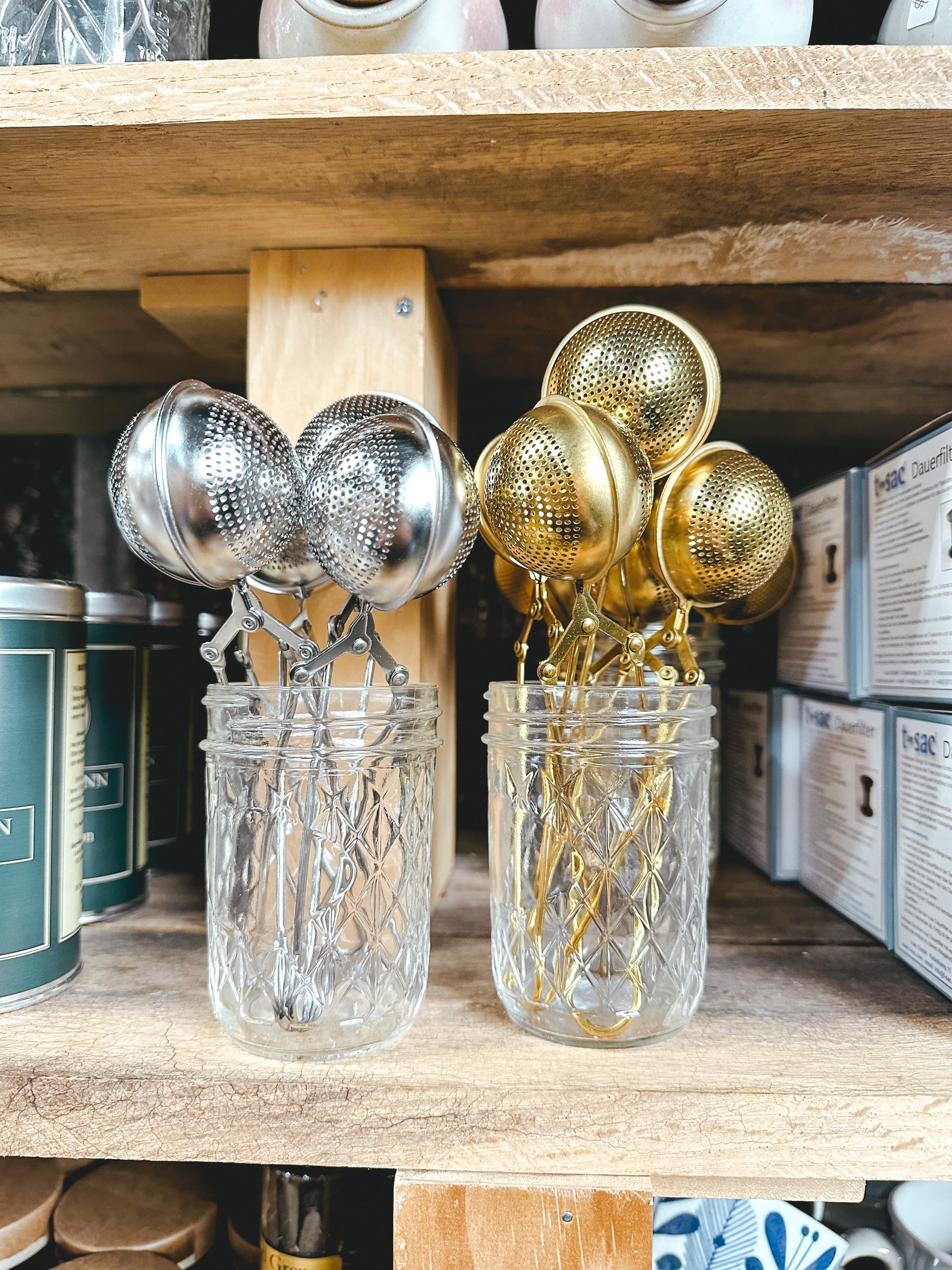 A collection of tea infuser balls with metallic parts, stored in a transparent jar on a wooden shelf, with other kitchen items in the background.