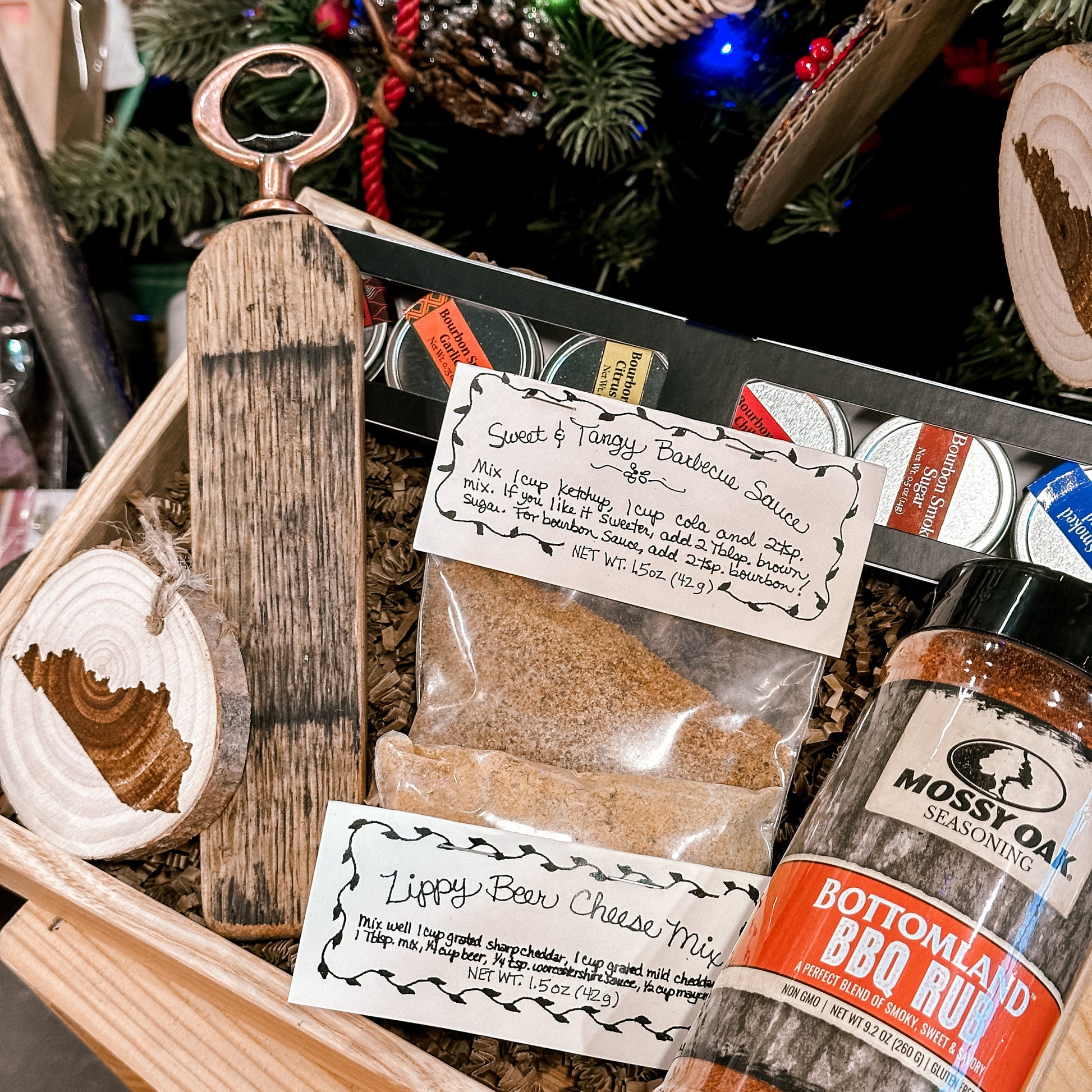 Gift basket with barbecue items including a bottle of Bottomland BBQ Rub, a wooden bottle opener, and a package of Lippy Bow Cheese Mix, set against a Christmas tree background.