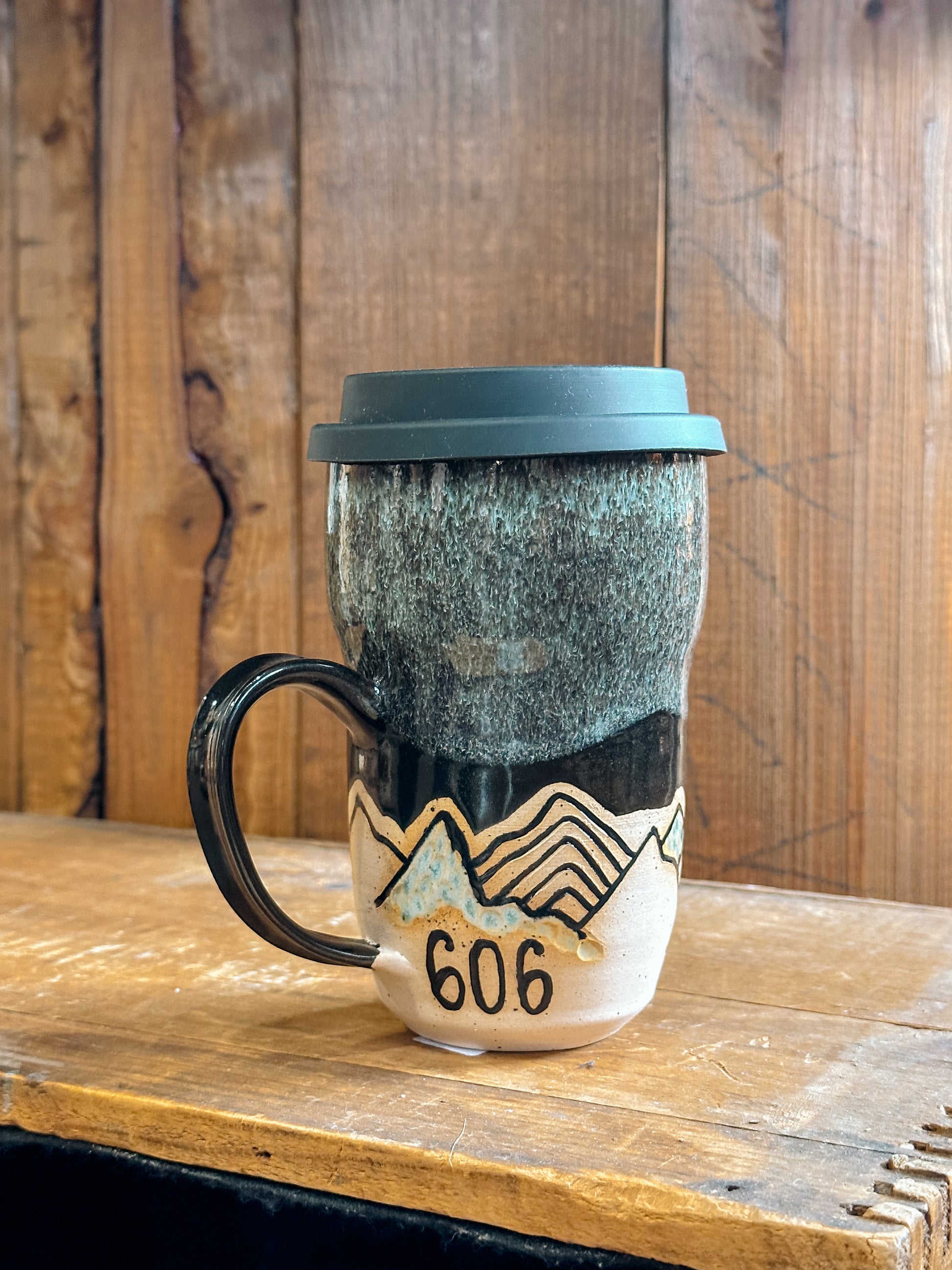 A handmade pottery travel mug with a silicone travel lid, featuring a cut-out design of Kentucky with landscapes, displayed on a wooden surface.