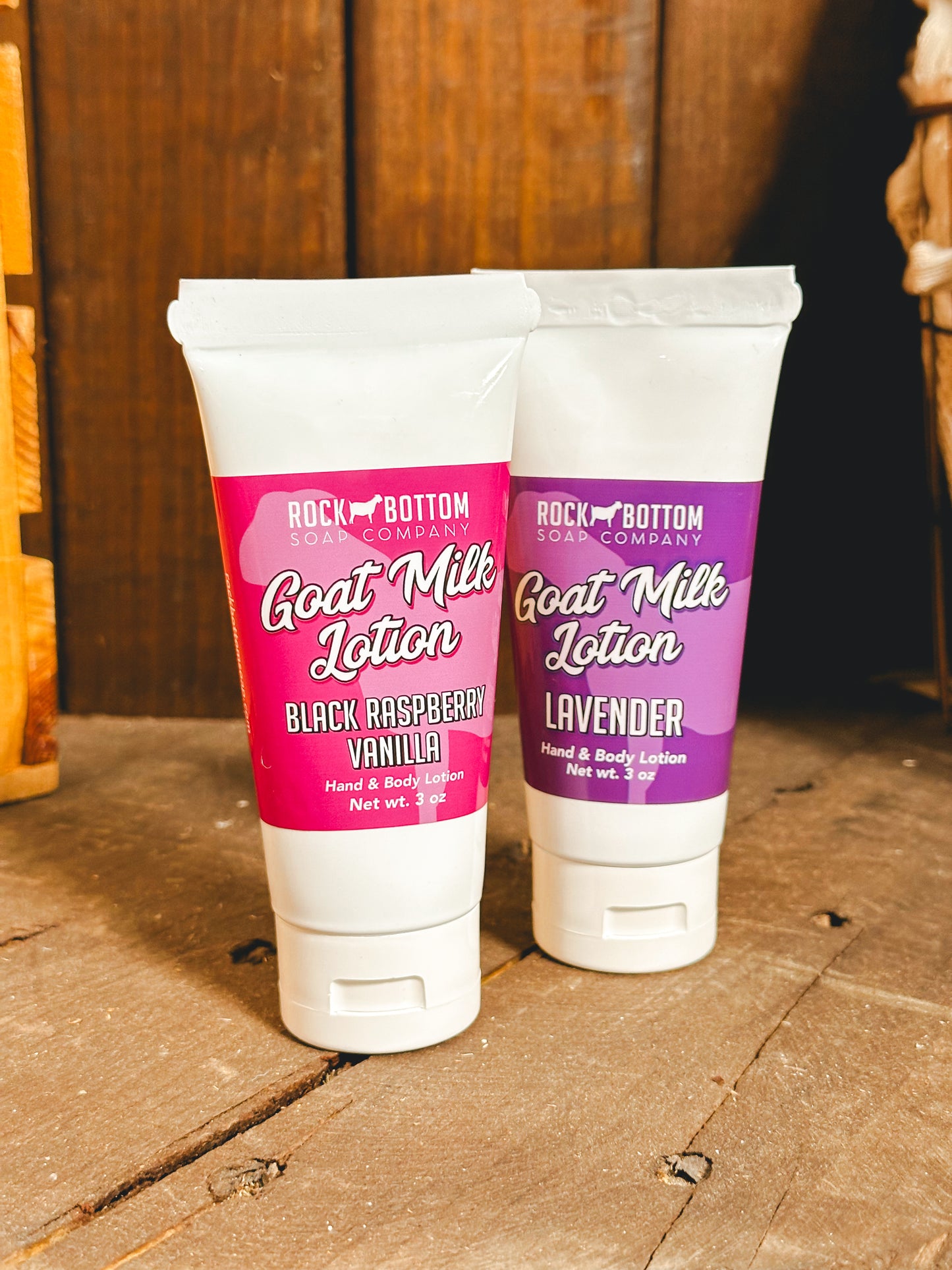 Two squeeze tubes of Goat Milk Lotion, one labeled 'Black Raspberry Vanilla' and the other 'Lavender', standing upright on a wooden surface.