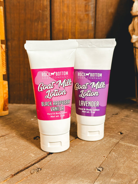 Two squeeze tubes of Goat Milk Lotion, one labeled 'Black Raspberry Vanilla' and the other 'Lavender', standing upright on a wooden surface.