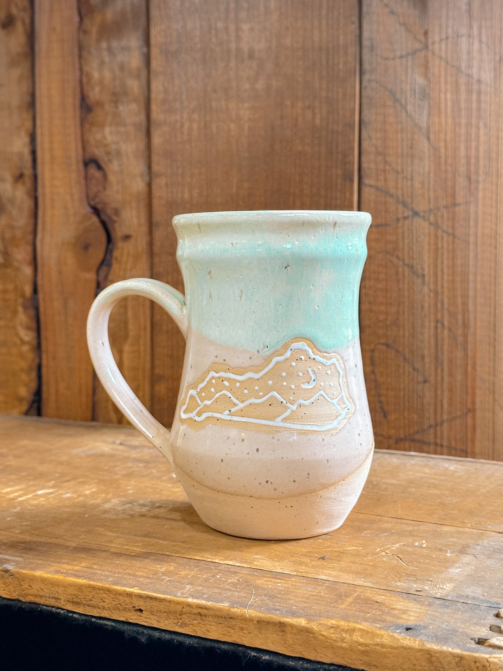 A handmade ceramic mug with a beige and green color scheme, featuring a cut-out design of Kentucky landscapes.
