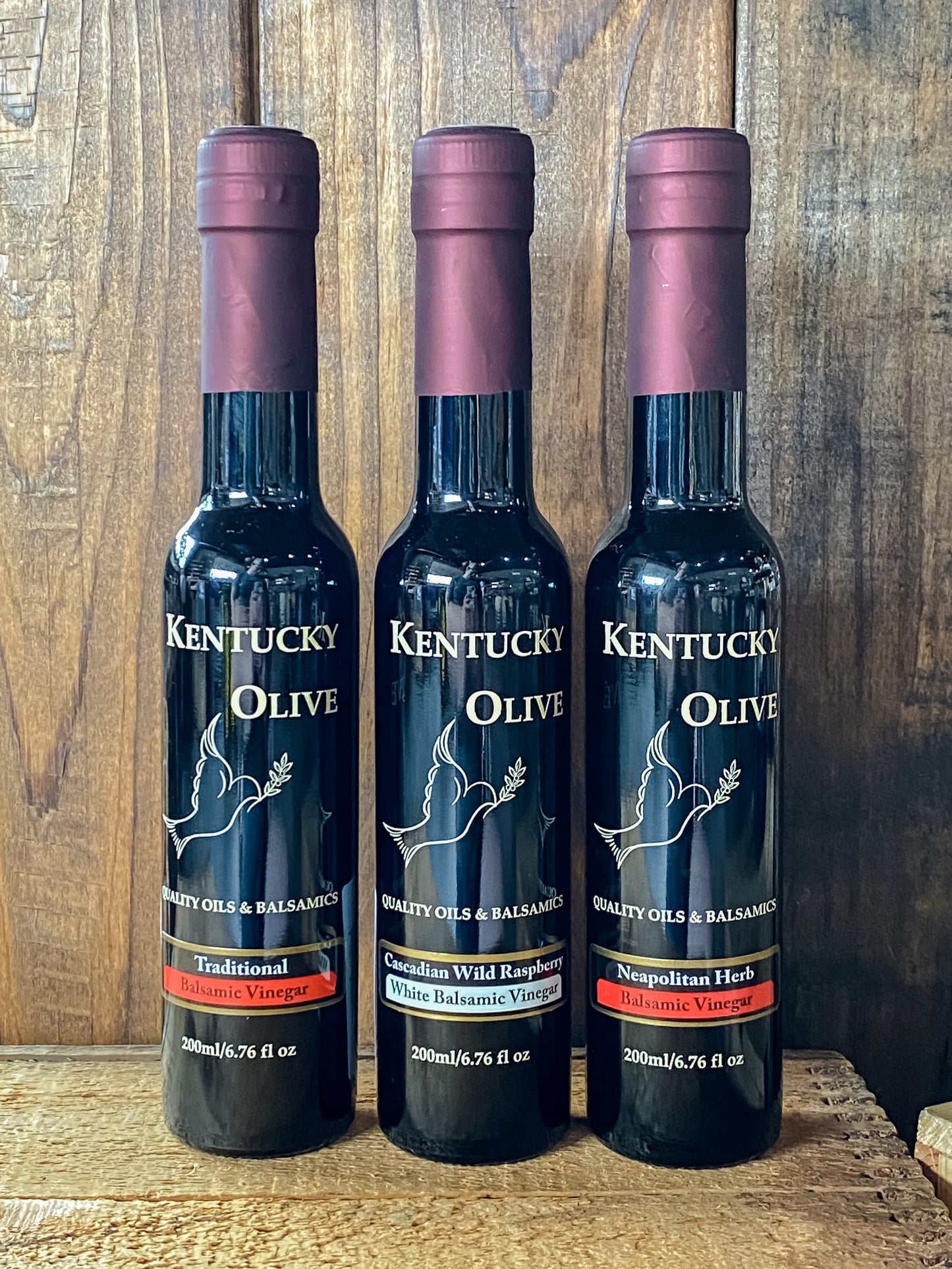 Three bottles of Kentucky Olive balsamic oil with dark labels and maroon caps, displayed against a wooden background.