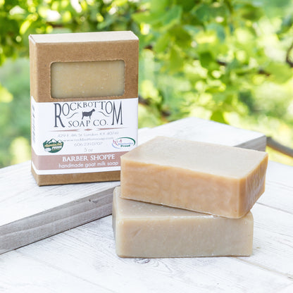 Masculine-scented goat milk soap bar on a handmade pottery soap dish with green foliage background – natural outdoor display