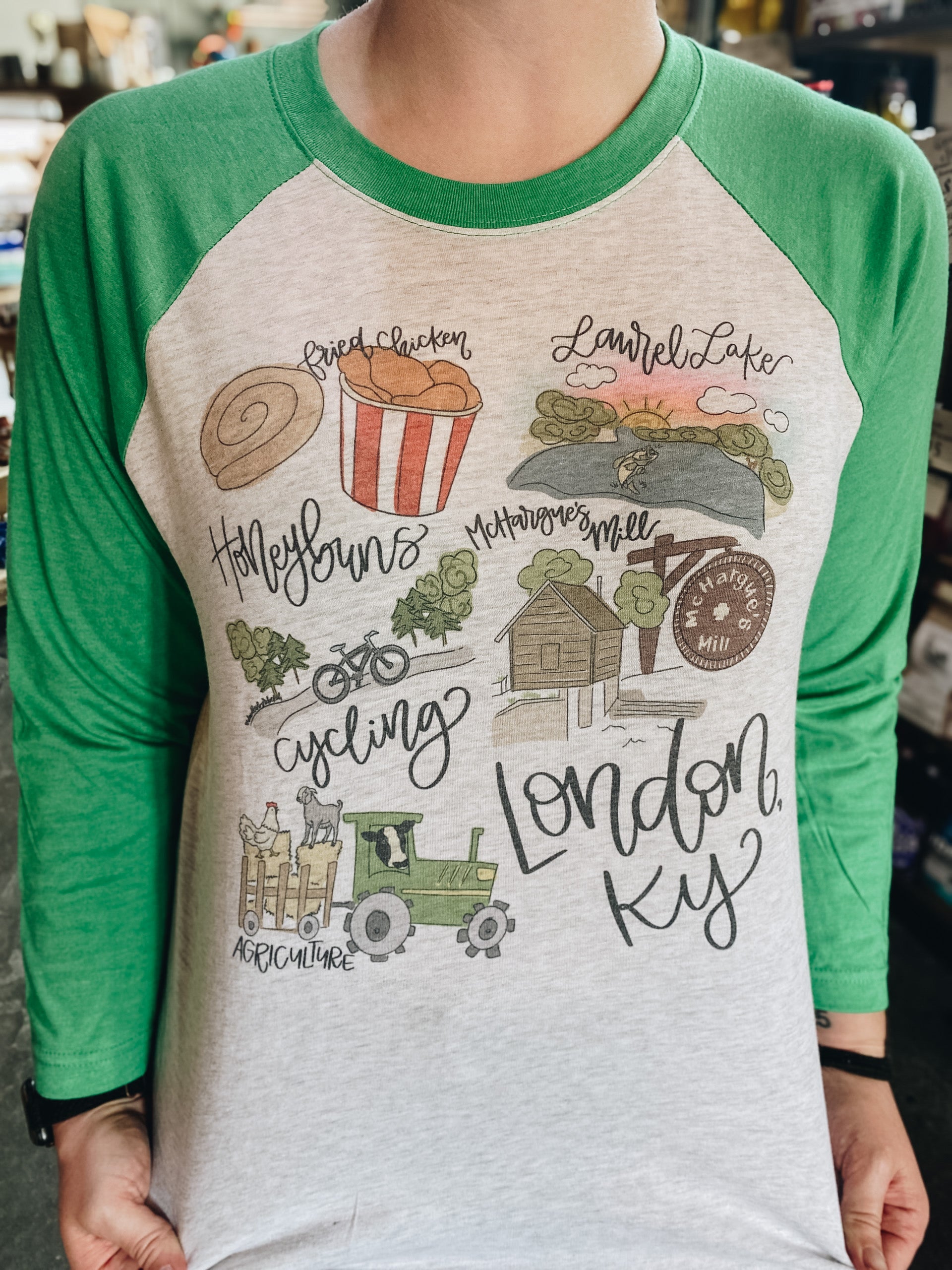 A person wearing a 'London, KY' raglan t-shirt with green sleeves and white body, featuring hand-illustrated elements related to London, Kentucky.